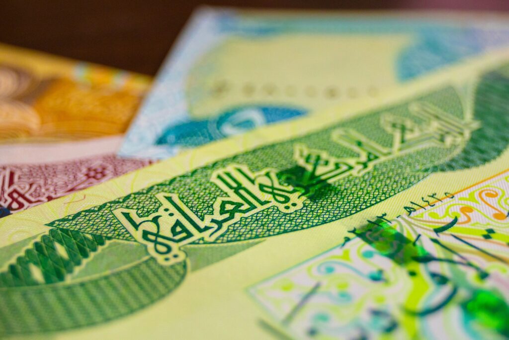 A close-up, shallow-depth-of-field shot of Iraqi Dinar banknotes. The foreground features green Arabic calligraphy and intricate patterns on a crisp bill, while other colorful notes blur into the background, highlighting the currency's detail.