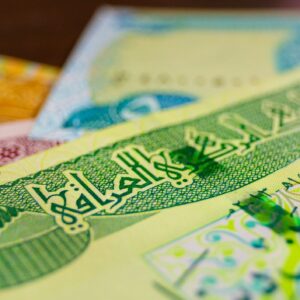 A close-up, shallow-depth-of-field shot of Iraqi Dinar banknotes. The foreground features green Arabic calligraphy and intricate patterns on a crisp bill, while other colorful notes blur into the background, highlighting the currency's detail.